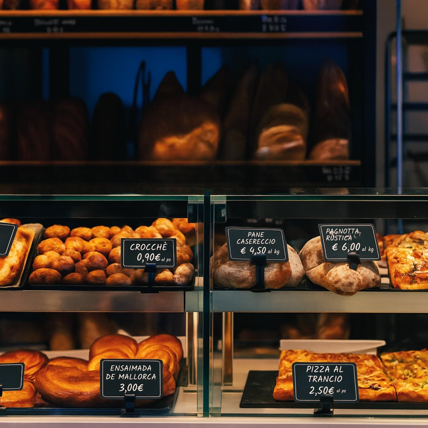 Cartellini neri esposti su banco panificio con pane, pizza e prodotti da forno